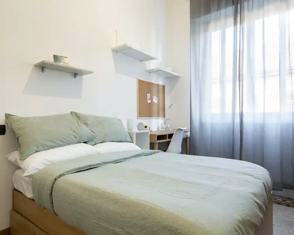 Bright, modern bedroom with a double bed dressed in soft green linens, a small desk by the window and minimalist shelving — ideal for a comfortable solo or couple stay.