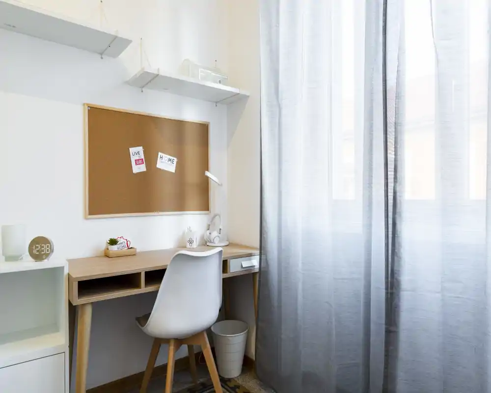 Bright, minimalist home office corner with a wooden desk, white chair, corkboard and soft natural light through sheer curtains — ideal for focused work or study.