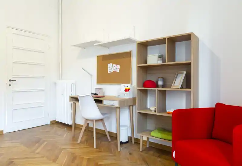 Bright, modern living area with a small desk, shelving unit and a vivid red armchair; wooden parquet and white walls create a clean, cozy atmosphere.