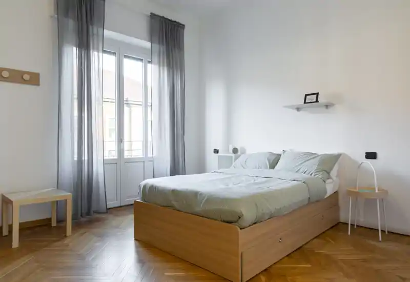 Camera da letto luminosa e minimalista con letto su pedana in legno, biancheria verde chiaro e ampia finestra che lascia entrare molta luce naturale. Comodini semplici e arredi neutri creano un'atmosfera calma e ariosa, ideale per un soggiorno riposante.