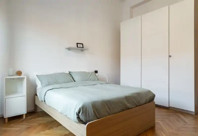 Camera da letto luminosa e minimalista con letto matrimoniale, comodino e grande armadio bianco; lenzuola verde tenue e pavimento in parquet caldo creano un'atmosfera moderna e rilassante.