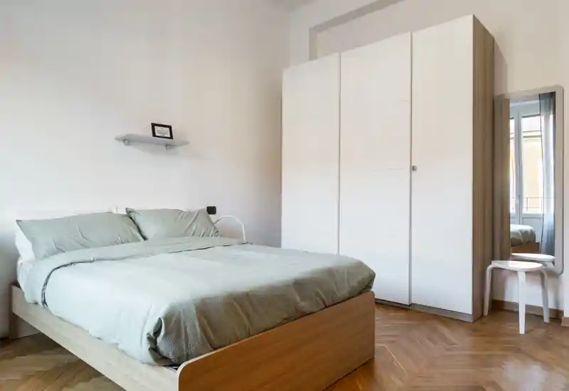 Camera da letto luminosa e minimal con grande letto in legno, biancheria verde salvia e armadio bianco; pavimento a spina di pesce caldo e luce naturale che creano un'atmosfera moderna e tranquilla.