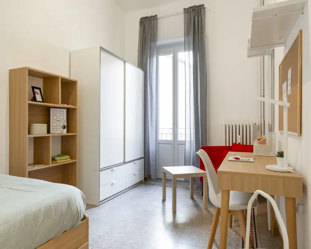 Bright, tidy bedroom with a visible single bed, wardrobe, shelving and a desk next to a window letting in natural light. The room features light wood furniture and a calm, modern decor ideal for a rental listing.