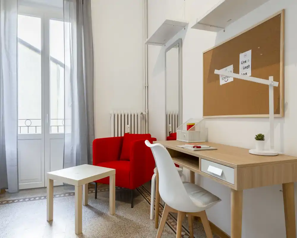Bright, stylish living area with a red armchair, small coffee table and a wooden desk by a tall window; modern, welcoming layout ideal for relaxing or working.