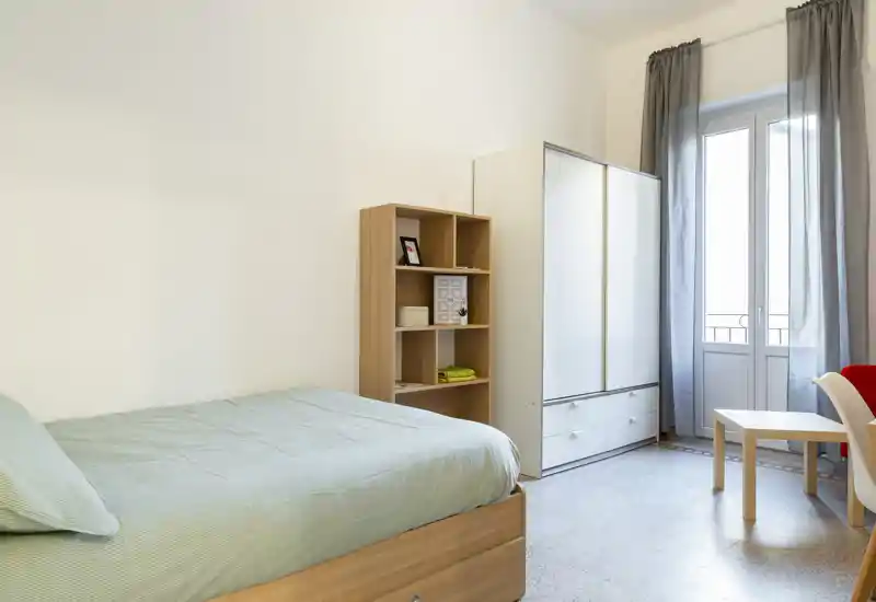 Dormitorio luminoso y sencillo con cama individual, armario y estantería junto a la ventana que deja entrar luz natural.