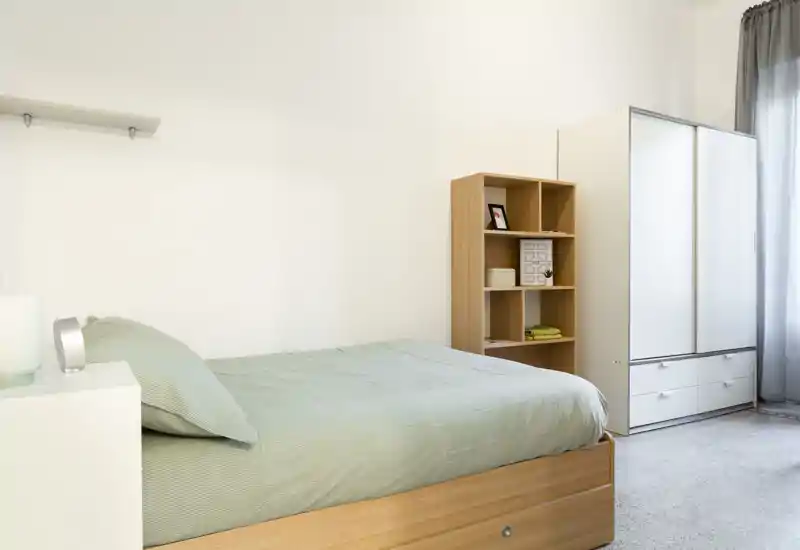 Habitación individual luminosa y minimalista con cama de madera, ropa de cama ordenada y armario junto a la ventana — ideal para un alquiler moderno y limpio.