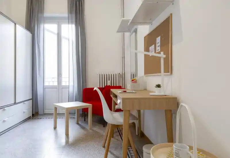 Rincón de habitación tipo estudio con escritorio, armario y pequeño espacio de estar. Mucha luz natural desde la ventana y mobiliario moderno y funcional.