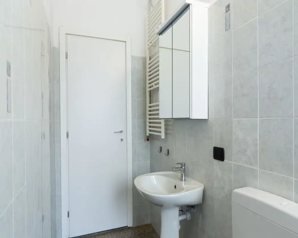 Compact, bright bathroom with a pedestal sink, mirrored cabinet and toilet. Pale gray wall tiles and a heated towel rail create a clean, minimalist look.