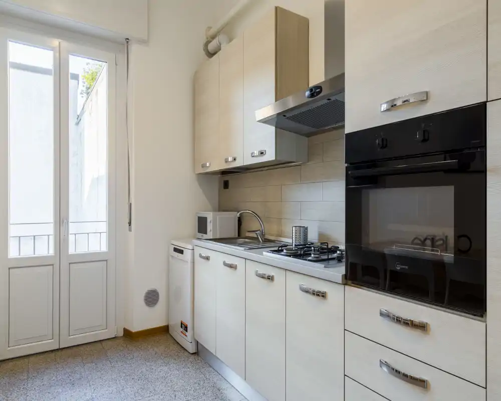 Modern compact kitchen with light wood cabinets, built-in oven, gas hob and sink; bright natural light from a tall glass door opens to a small balcony or exterior space.