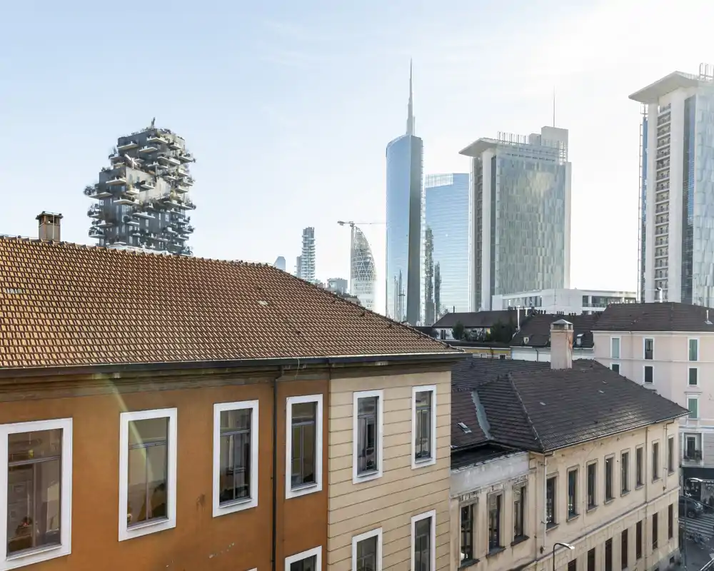 Vista cittadina da una finestra o balcone con palazzi storici in primo piano e grattacieli moderni sullo sfondo, luce diurna chiara e cielo azzurro.