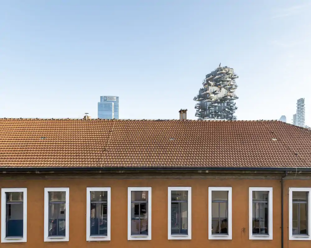 Vista esterna del tetto con tegole arancioni e della facciata dell'edificio con diverse finestre; sullo sfondo si intravedono moderni grattacieli sotto un cielo azzurro limpido.