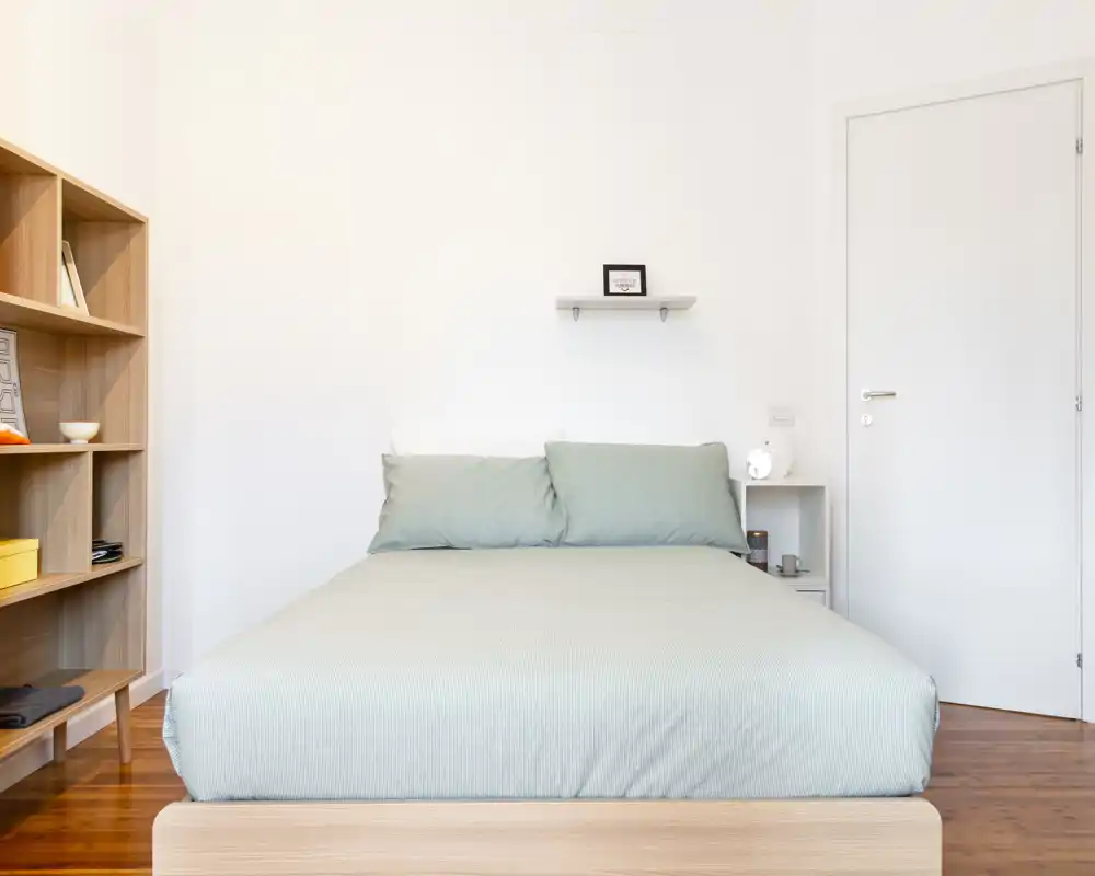 Camera da letto luminosa e minimal con letto matrimoniale centrale su parete bianca, biancheria verde tenue e pavimento in legno. Scaffalatura semplice e comodino creano un aspetto moderno e tranquillo, ideale per annunci.
