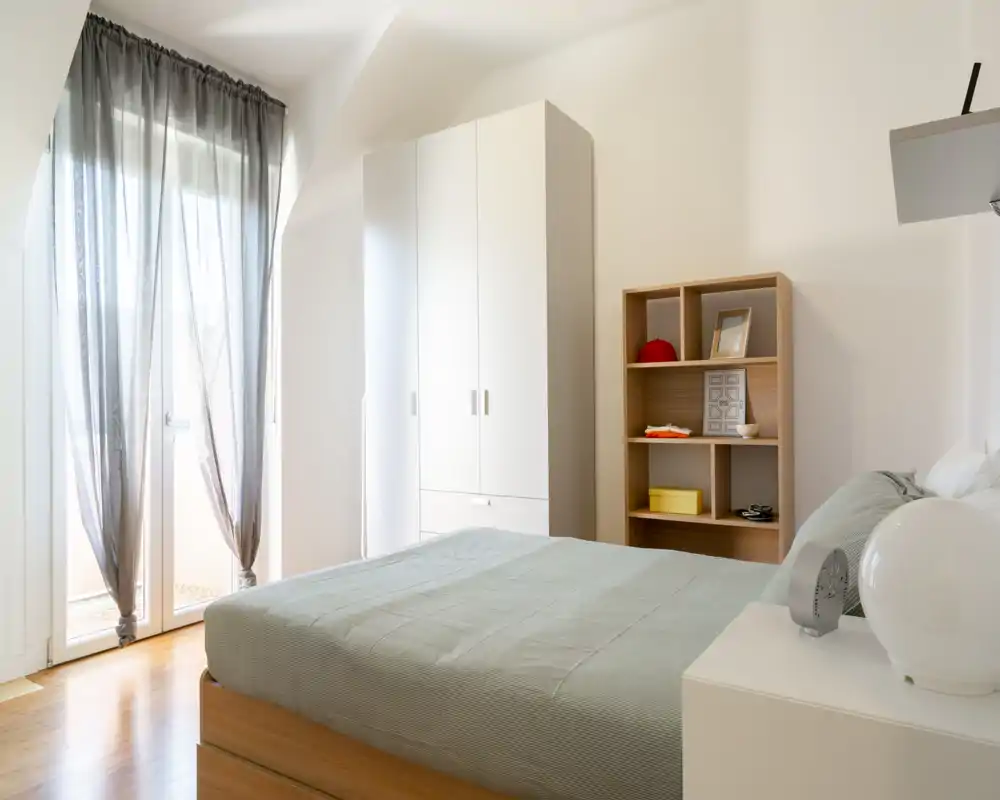 Camera da letto moderna e luminosa con letto matrimoniale ben rifatto, armadio e scaffale. Ampie porte-finestre con tende leggere che lasciano entrare molta luce naturale.