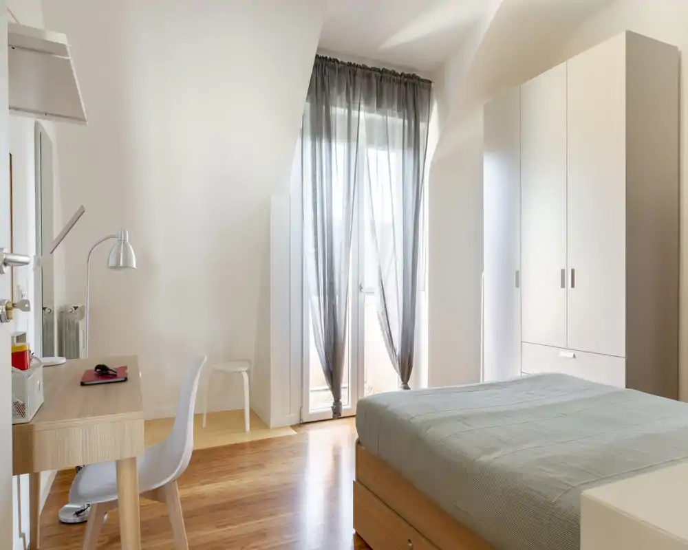 Camera da letto moderna e luminosa con letto matrimoniale, armadio e una scrivania vicino alla porta del balcone che lascia entrare molta luce naturale.