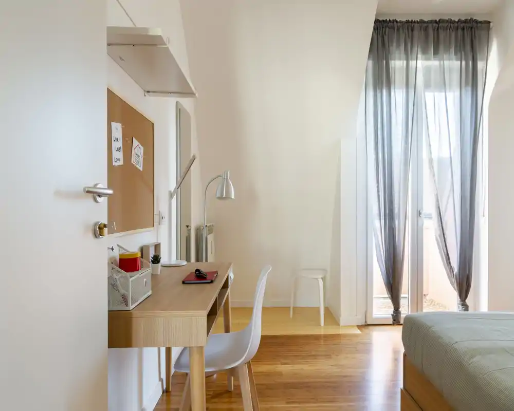 Camera da letto luminosa e minimal con scrivania in legno e un letto comodo accanto alla porta del balcone: ideale per soggiorni rilassanti e lavoro da remoto.