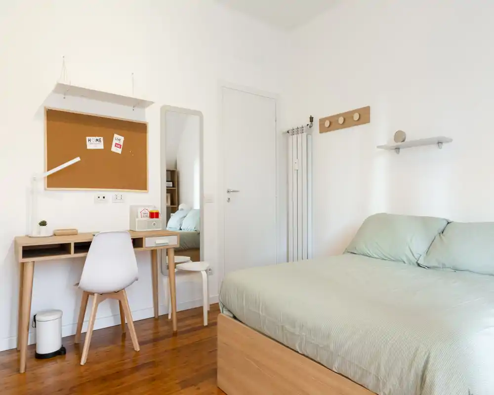 Camera da letto luminosa e minimalista con un grande letto con biancheria verde chiaro, una scrivania in legno e uno specchio sulla parete opposta — stile moderno e pavimenti in legno caldi.