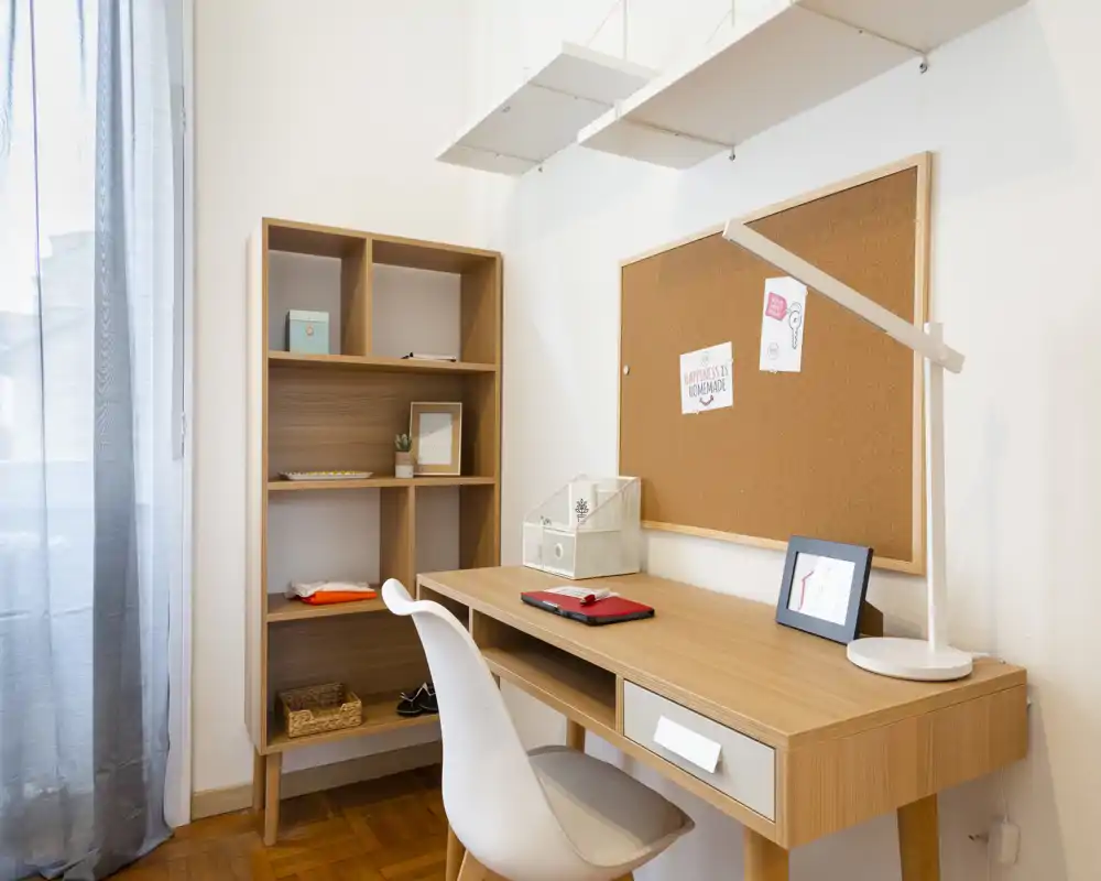 Espace de travail lumineux et soigné avec bureau en bois, chaise blanche et panneau en liège — idéal pour étudier ou télétravailler. La lumière naturelle filtre à travers un rideau fin et les étagères apportent du rangement pratique.