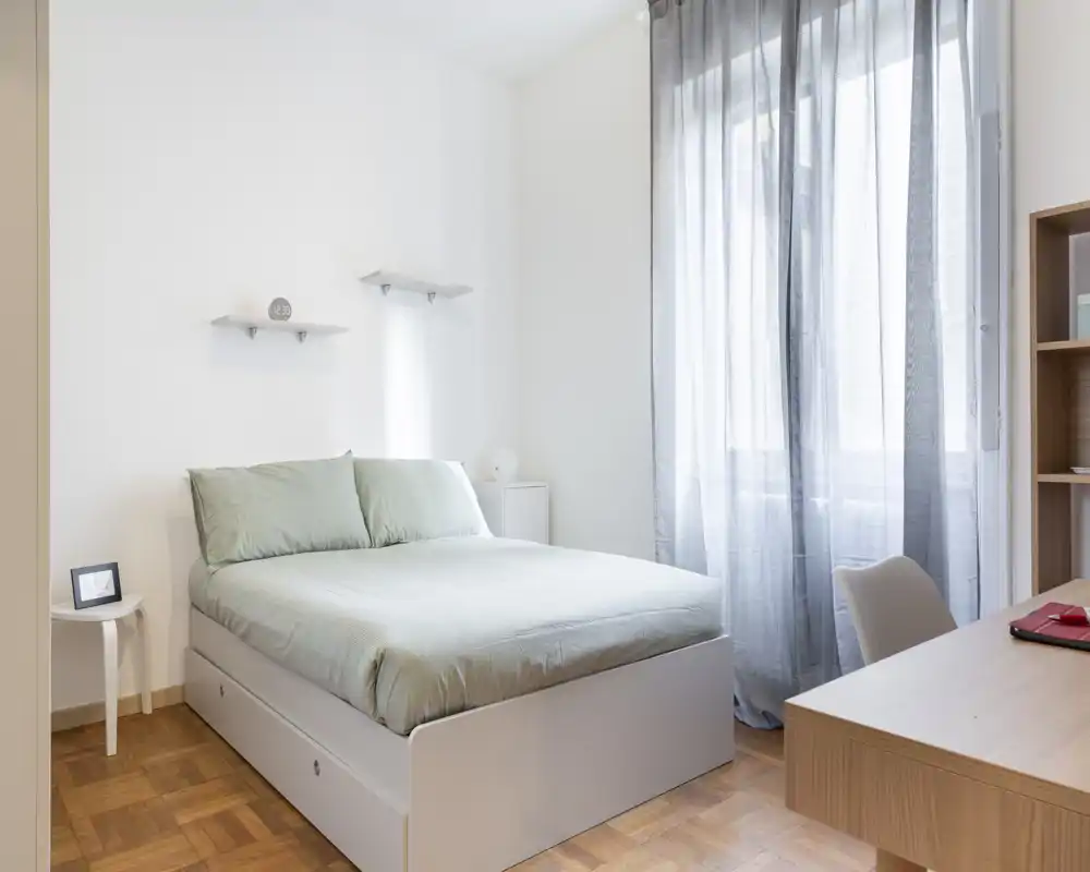Chambre lumineuse et minimaliste avec un lit double, linge de lit vert pâle, lumière naturelle venant d'une grande fenêtre et un petit bureau — idéale pour un séjour calme.