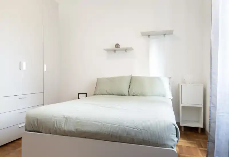 Chambre lumineuse et minimaliste avec un lit double habillé de linge vert pâle, une armoire blanche et une petite table de chevet — idéale pour un séjour calme et moderne.