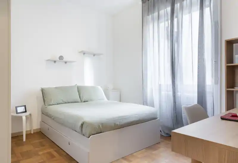 Chambre lumineuse et minimaliste avec un lit double, linge de lit vert pâle, lumière naturelle venant d'une grande fenêtre et un petit bureau — idéale pour un séjour calme.