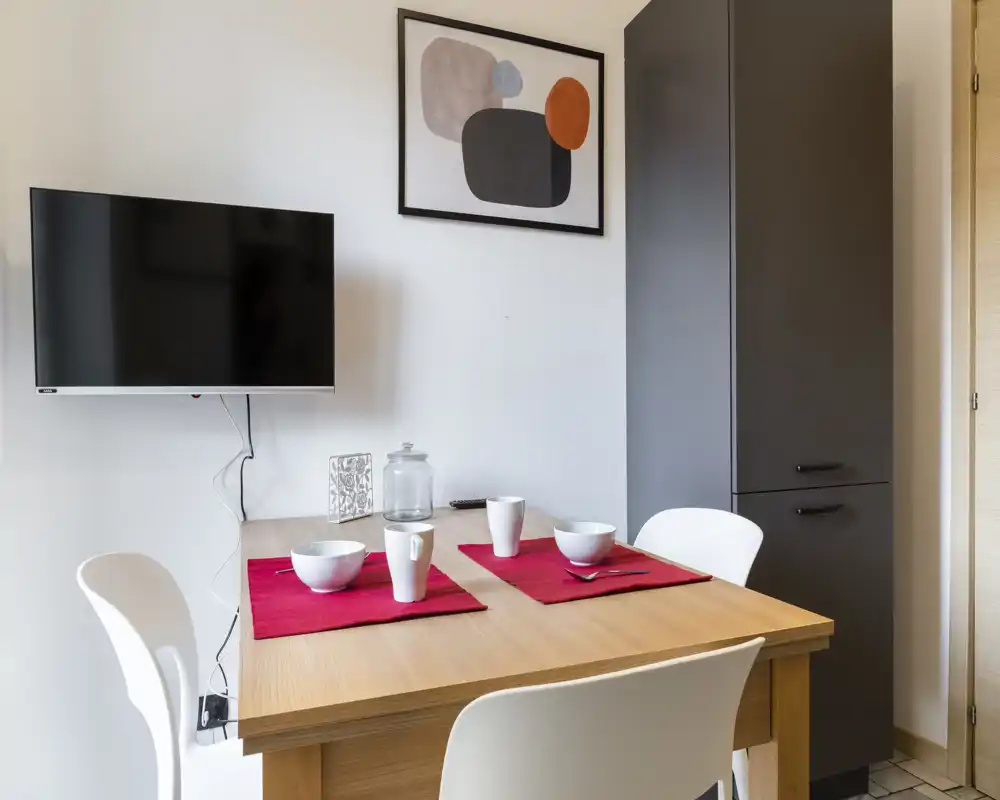 Coin repas compact avec table en bois dressée pour deux et chaises blanches modernes, mis en valeur par des sets de table rouges, un grand meuble de rangement sombre et une œuvre abstraite encadrée — lumineux et soigné pour un petit appartement.