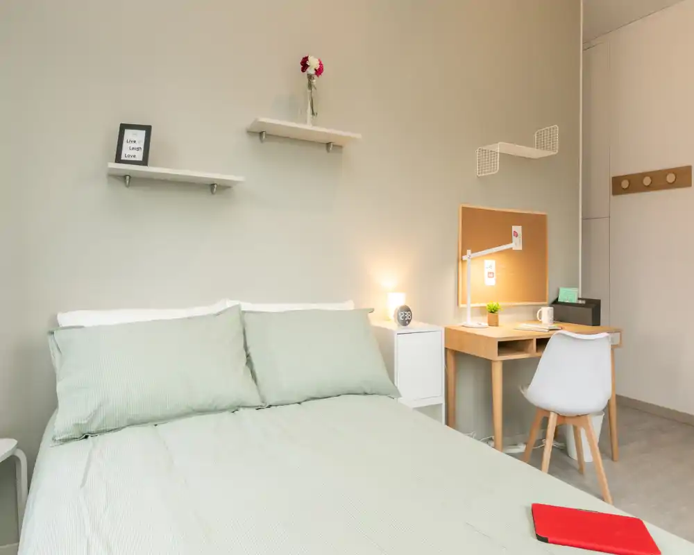 Bright, neatly staged bedroom with a double bed dressed in soft green linens, simple wall shelves and a compact desk workspace — ideal for a cozy rental stay.