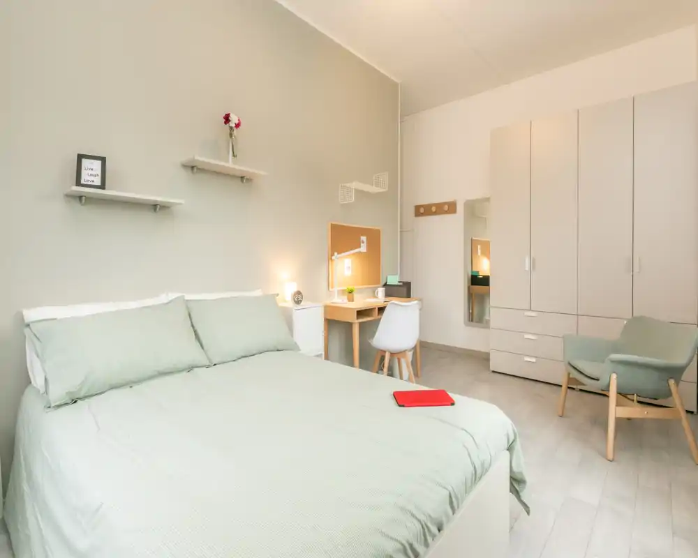 Bright, modern bedroom with a neatly made double bed, a small desk and chair, wardrobe and a comfortable armchair — tastefully decorated in soft green and neutral tones.
