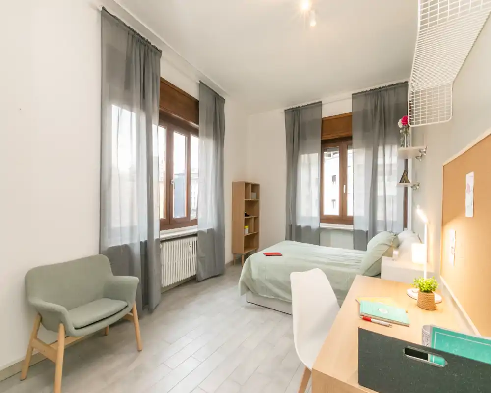 Bright, well-staged bedroom with a comfortable double bed, desk area and armchair. Large windows with sheer curtains let in plenty of natural light for a pleasant, airy atmosphere.