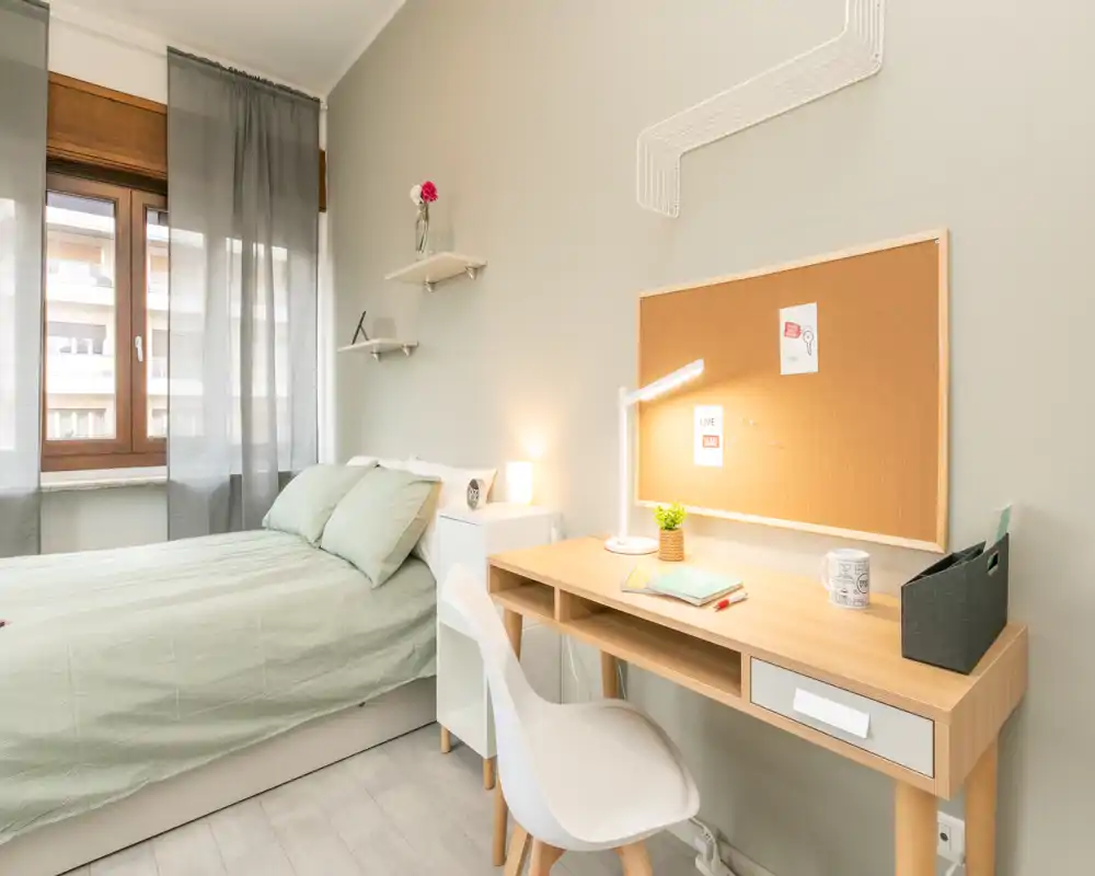 Bright, well-staged bedroom with a full bed, bedside table and a practical desk under a corkboard — ideal for working and relaxing.