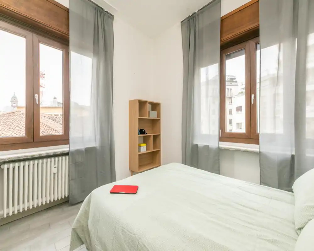 Bright, simply furnished bedroom with a double bed positioned beneath two large windows, light-green bedding and a small wooden shelving unit; ideal for a calm, sunlit stay.