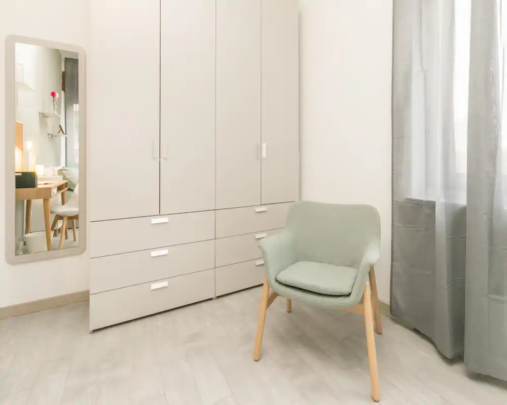 Bright, minimal bedroom corner with a large built-in wardrobe, a light-green armchair and a full-length mirror reflecting part of a bed. Neutral tones and soft daylight create a calm, modern atmosphere.