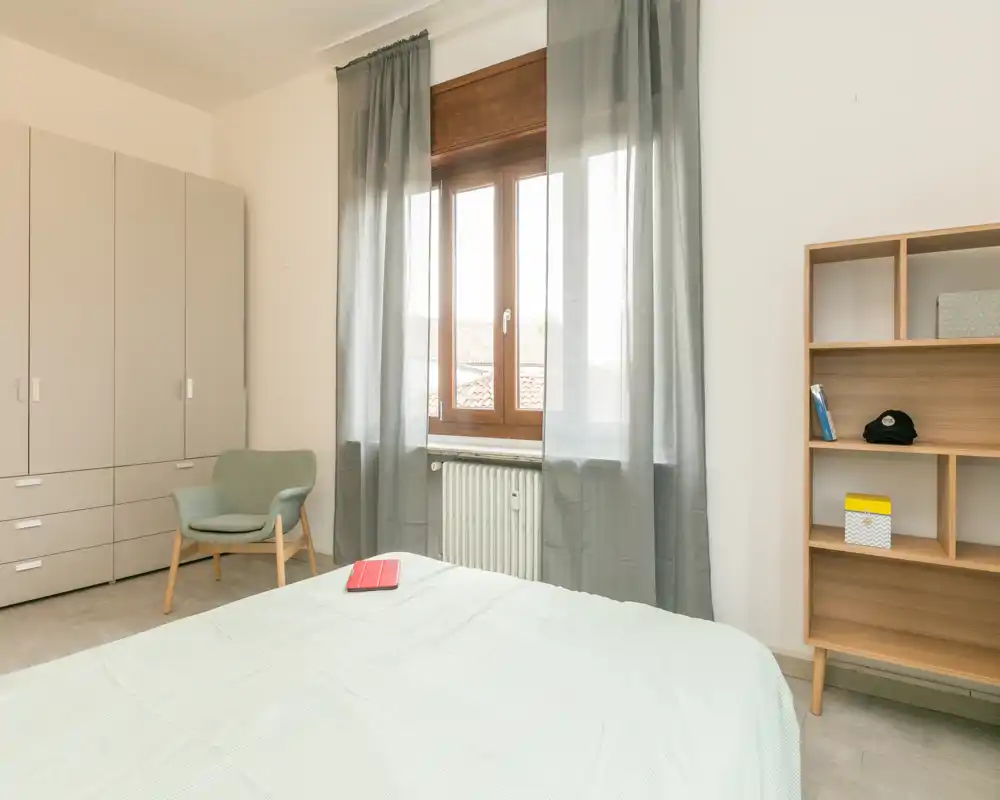 Bright, minimalist bedroom with a large bed in the foreground, a wardrobe, an armchair and a shelving unit by the window letting in natural light.