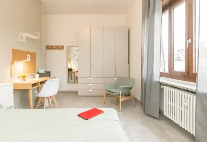 Bright, modern bedroom with a neatly made bed in the foreground, a wooden desk and chair, wardrobe and a lounge chair near a large window letting in abundant natural light.