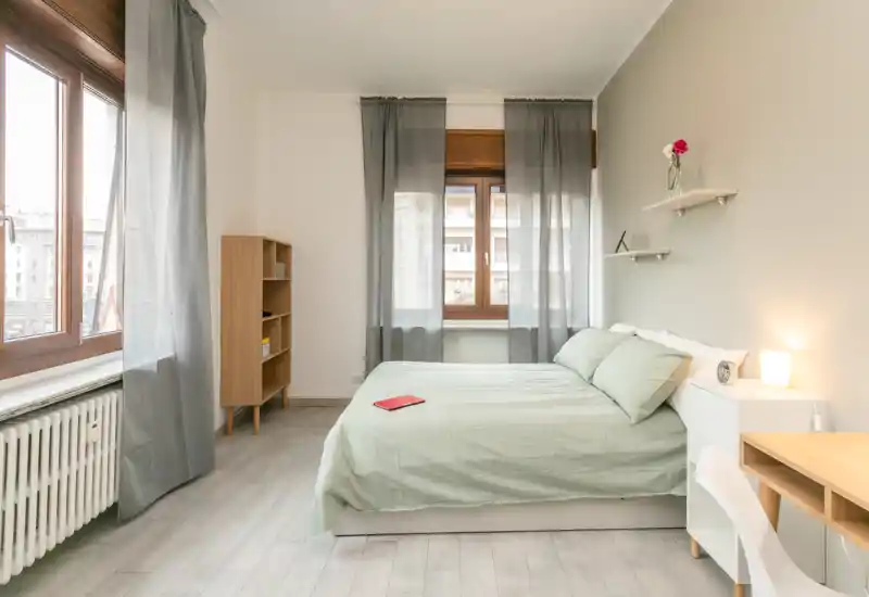 Bright, modern bedroom with a double bed dressed in light linens, large windows with sheer curtains, a small shelf with decor and a bedside lamp creating a calm, airy atmosphere.