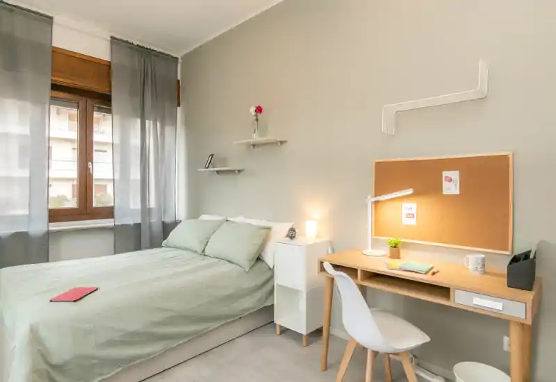 Bright, tidy bedroom with a double bed dressed in soft green linens, a bedside cabinet and a study desk with cork board — ideal for a student or professional.