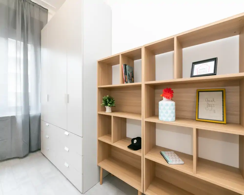 Area di deposito ordinata con scaffalature in legno chiaro e un armadio bianco accanto a una finestra con tenda. Oggetti decorativi e libri sono disposti sugli scaffali aperti, creando un aspetto minimale e ordinato.