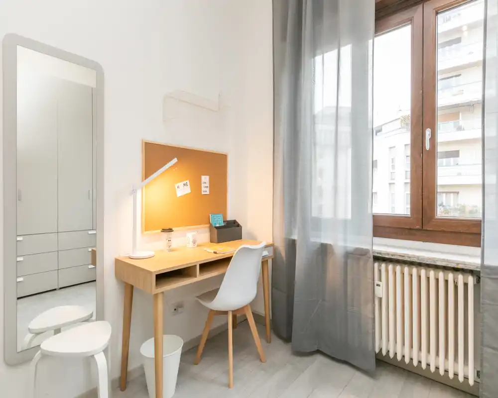 Angolo camera luminoso e minimal con scrivania in legno, sedia e bacheca vicino a una grande finestra che lascia entrare luce naturale.