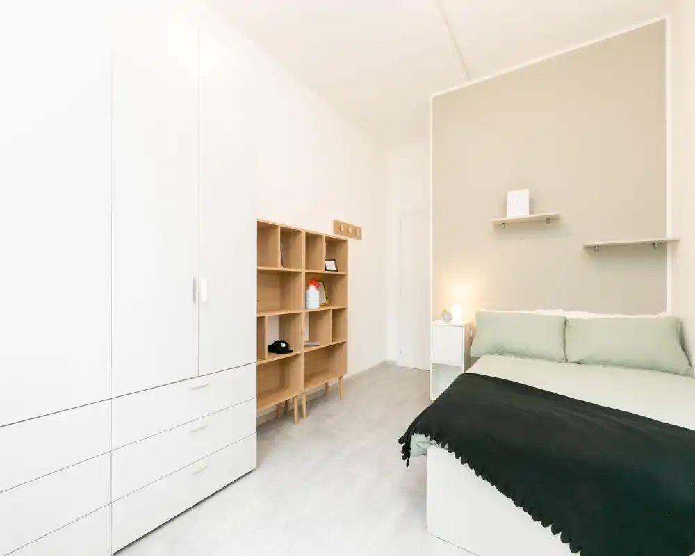 Camera da letto luminosa e minimal con letto matrimoniale rifatto, comodino e un armadio alto. Toni neutri e scaffalature in legno chiaro creano un'atmosfera moderna e rilassante.