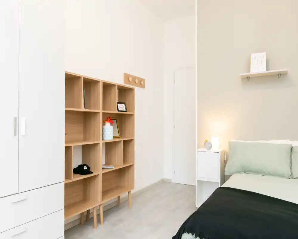 Camera da letto luminosa e minimal con letto rifatto, comodino e scaffalatura aperta — ambiente moderno e delicatamente arredato, ideale per un annuncio.