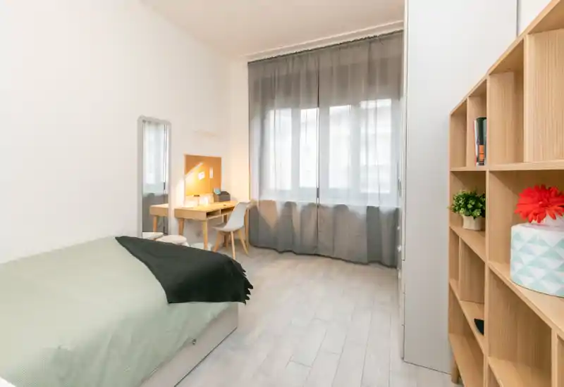 Camera singola luminosa e moderna con letto comodo, scrivania con lampada e scaffalatura aperta. Toni neutri e luce naturale morbida ideali per un annuncio di affitto.