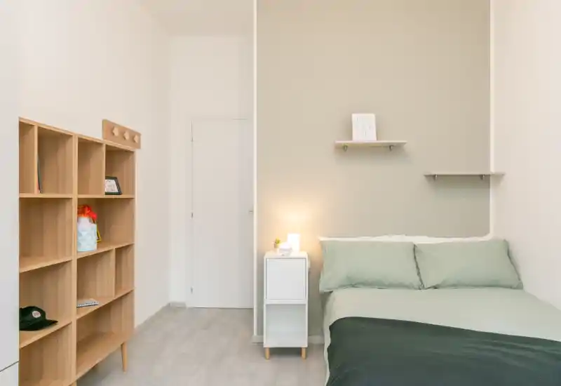 Camera da letto luminosa e minimal con letto ben rifatto, comodino con lampada e libreria in legno — toni verdi e neutri che creano un'atmosfera rilassante.