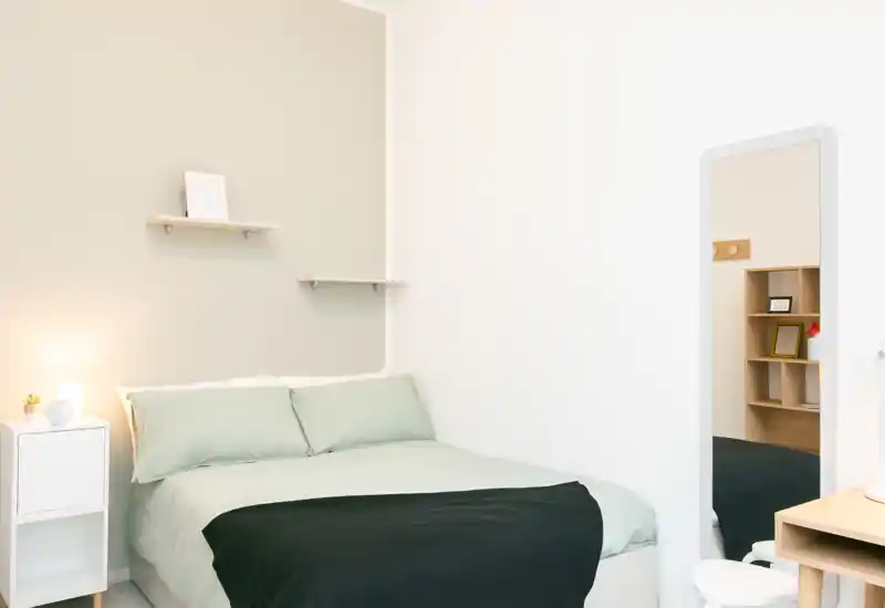 Camera da letto luminosa e minimal con letto matrimoniale ben rifatto, comodino con lampada e specchio intero che aggiunge profondità.
