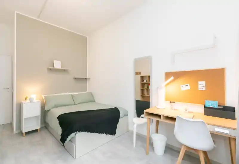 Camera da letto moderna e luminosa con letto matrimoniale ben rifatto, comodino con lampada, specchio a figura intera e scrivania con pannello in sughero — ideale per uno soggiorno confortevole o per studenti.