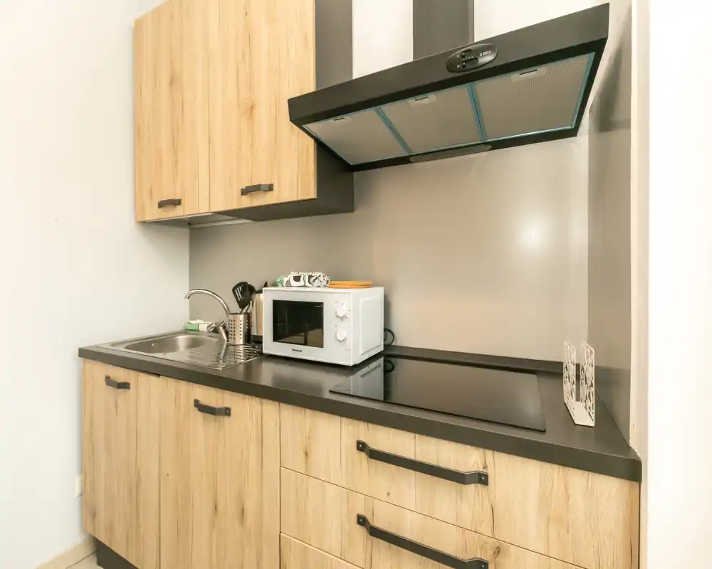 Compact modern kitchenette with wood-effect cabinets, electric cooktop, stainless sink and a countertop microwave — bright, clean and well organized for short-stay rentals.