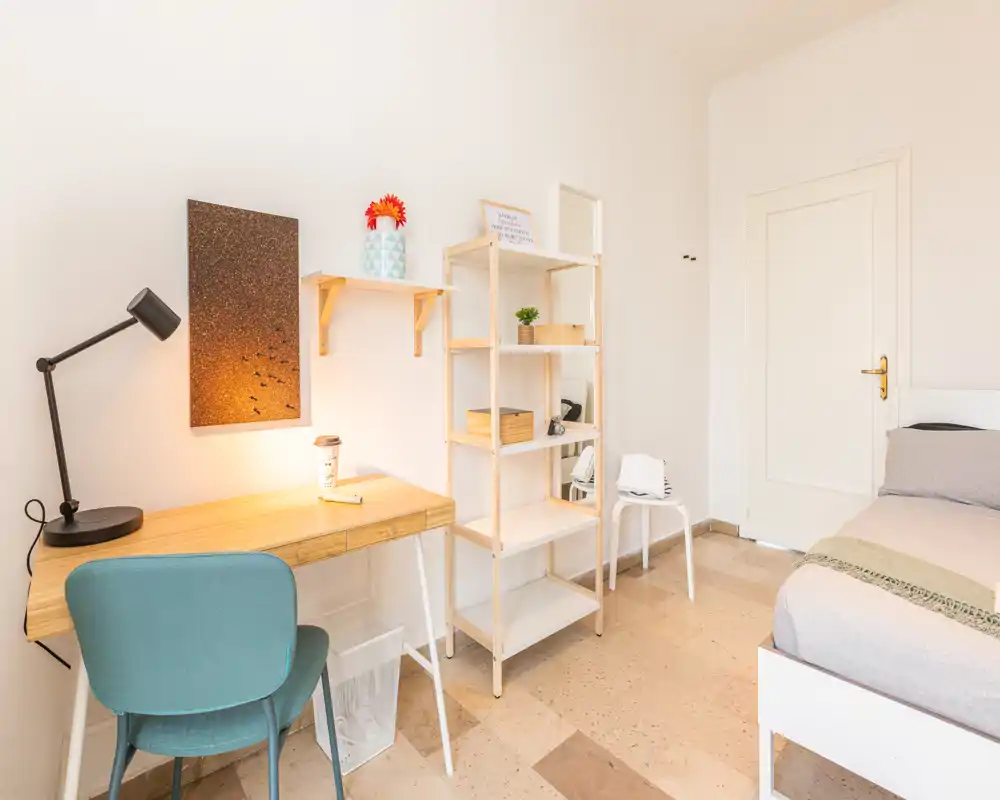 Habitación luminosa y ordenada con cama individual, escritorio de madera y estantería, ideal para un estudiante o persona sola. Iluminación cálida y mobiliario moderno y sencillo.