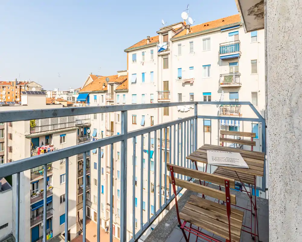 Pequeño balcón soleado con barandillas metálicas, dos sillas plegables de madera y una mesa a juego con vistas a los edificios vecinos — ideal para el café matutino.