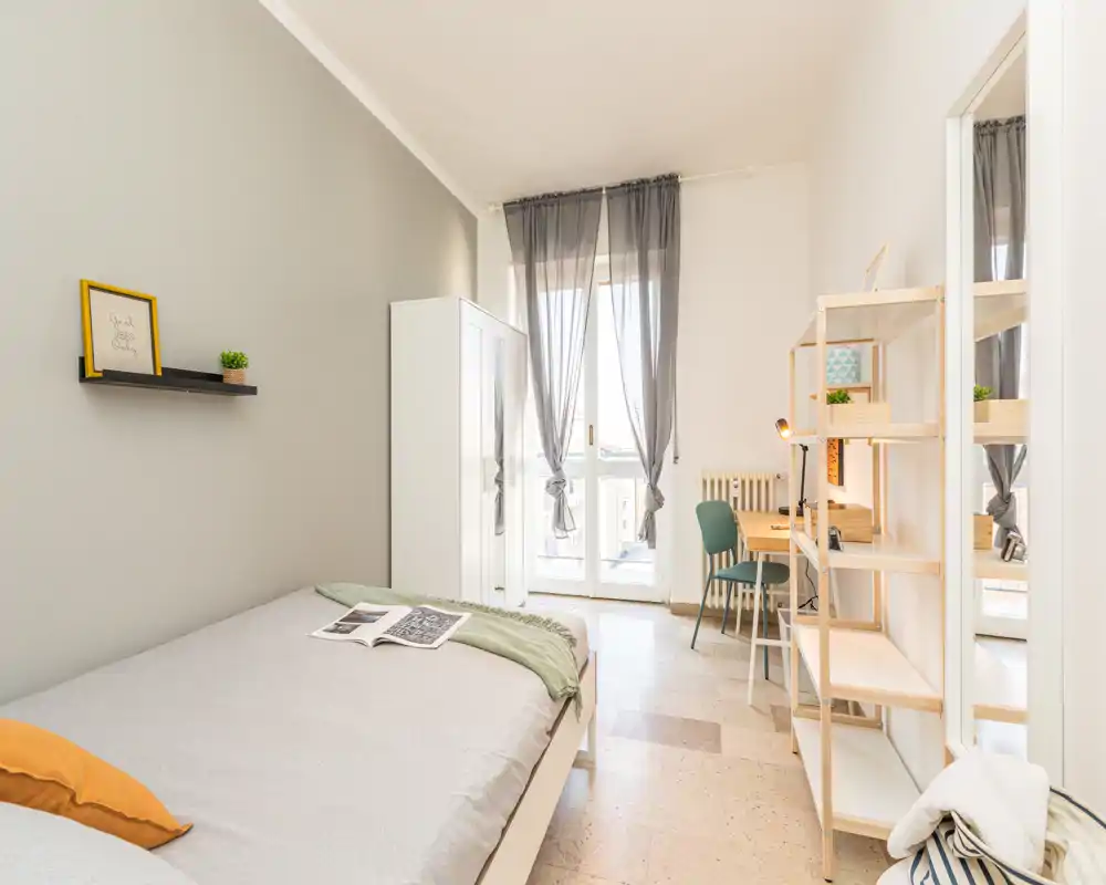 Dormitorio moderno y luminoso con cama doble, armario y un escritorio junto a la puerta del balcón; tonos neutros y textiles suaves que crean un ambiente acogedor.