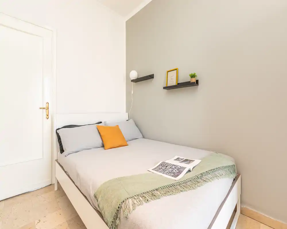 Dormitorio luminoso y minimalista con cama doble con ropa de cama neutra, una manta verde y un cojín amarillo; estantes flotantes y luz suave crean un ambiente moderno y tranquilo.