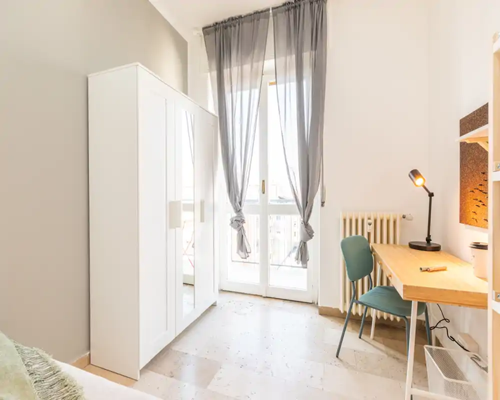 Dormitorio luminoso y minimalista con armario blanco, escritorio y puerta al balcón que aporta luz natural. Se ve parcialmente la cama en el borde izquierdo; hay un escritorio de madera y una silla junto al radiador.