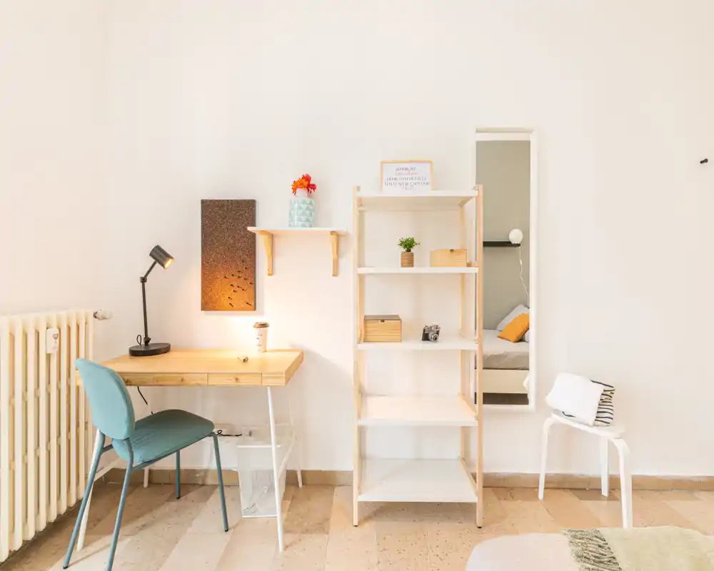 Rincón de dormitorio luminoso y minimalista con escritorio pequeño y estantería. Luz natural, textiles suaves y esquina de la cama visible, perfecto para un estudio acogedor.